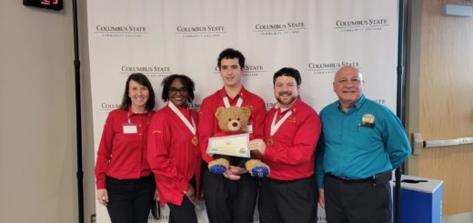 Phi Theta Kappans standing in front of a photo board presenting the Howdy Bear Certificate of Spirit and a stuffed teddy bear to go with it.