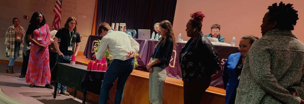 10 Phi Theta Kappans lined up on opposite sides of a table, 5 in each line. The ones at the front of both lines stack disposable cups in a pyramid formation.