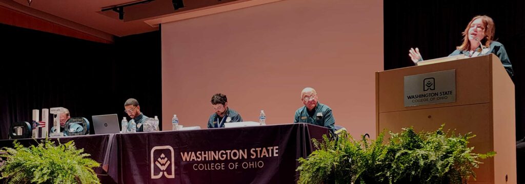 5 Regional Officers on the conference stage. One is stood speaking at a lectern while the other four are seated at tables facing the audience.