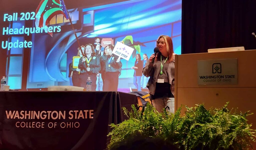 PTK Headquarters representative on the conference stage holding a handheld microphone with a powerpoint displayed in the background stating "Fall 2024 Headquarters Update".